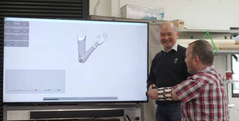 Two men are interacting with a large screen displaying a 3D model of a prosthetic arm. One, wearing a partial prosthetic arm, gestures while the other smiles and observes. The setting appears to be a laboratory or workshop filled with various equipment in the background, clearly depicting an innovative workline.