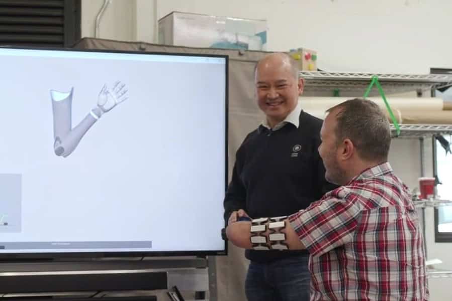 Two men are interacting with a large screen displaying a 3D model of a prosthetic arm. One, wearing a partial prosthetic arm, gestures while the other smiles and observes. The setting appears to be a laboratory or workshop filled with various equipment in the background, clearly depicting an innovative workline.